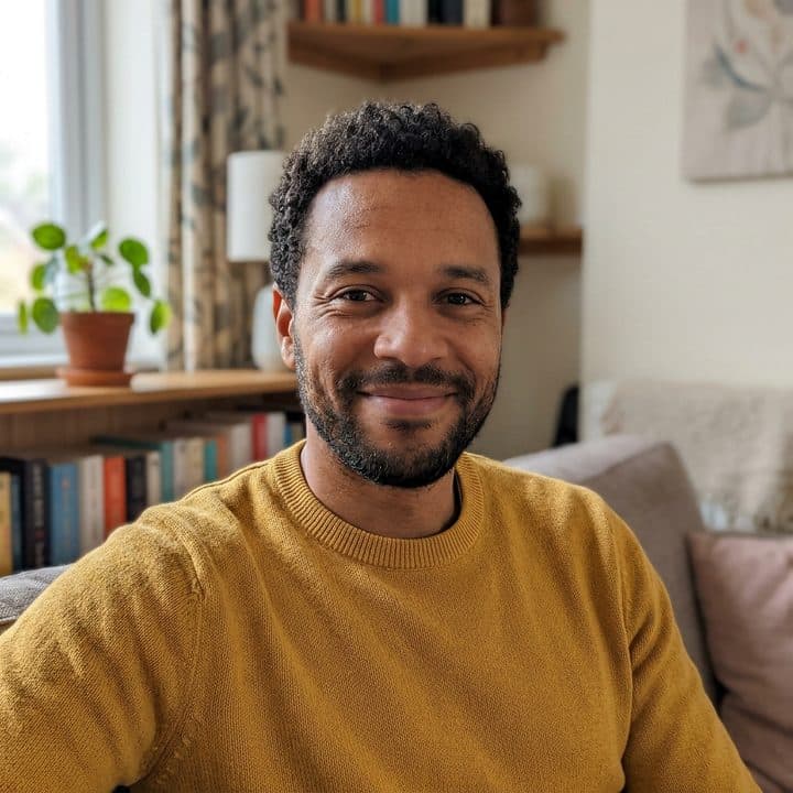 A photograph of a smiling man with short curly hair and a trimmed beard, in a mustard-yellow sweater.